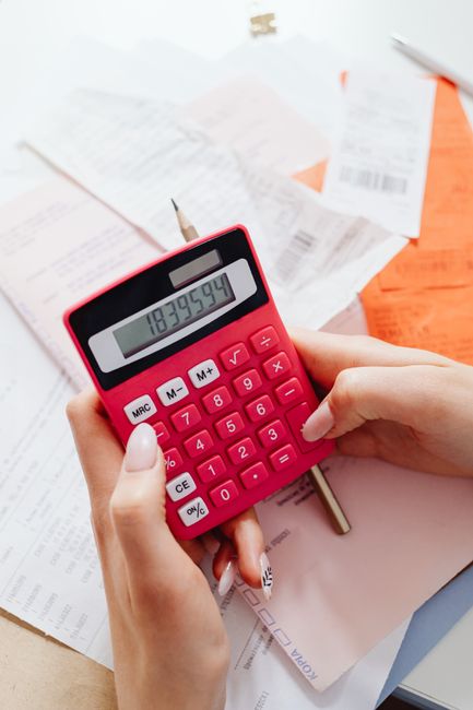 Close-up of a person's hands holding a pink calculator displaying the number 187,395.94, with a silver pen resting on a stack of papers and documents on a wooden table. In the background, there are ga
