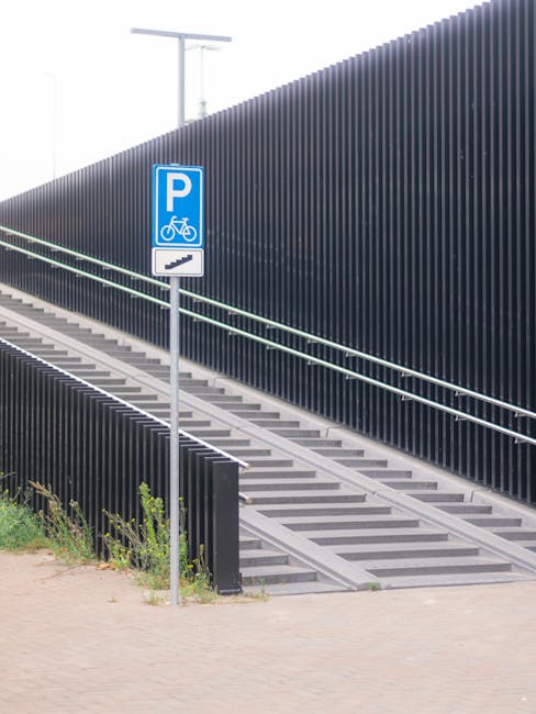 A concrete outdoor slope with integrated stairs leading up to a raised platform, accompanied by a metal handrail along the ramp and stairs for accessibility. To the left, a blue parking sign indicates